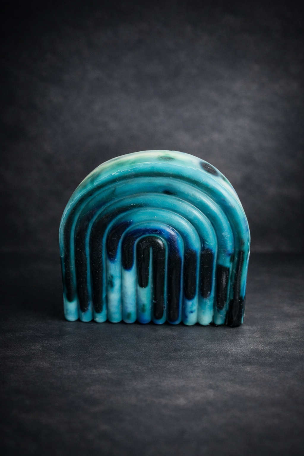 Teal and deep blue arch-shaped wax sculpture with layered curved ridges, photographed on a dark moody studio background.