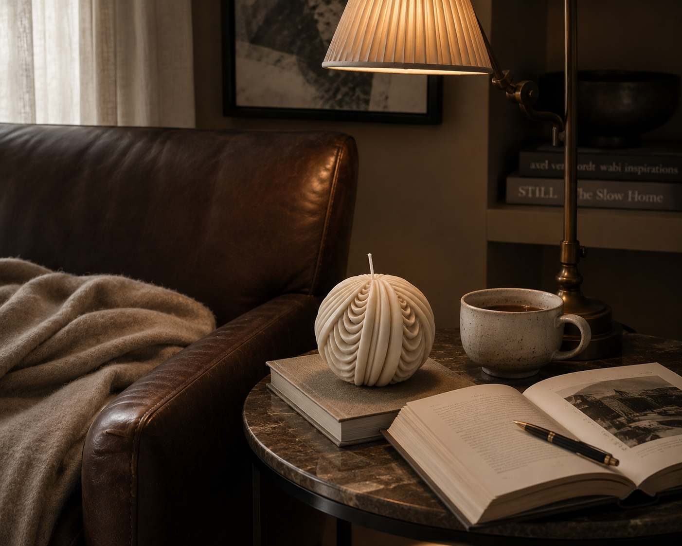 Cashmere Obsidian Sphere candle displayed on stacked books atop a marble side table, paired with a ceramic mug and open book beneath warm ambient lighting in a refined living space.