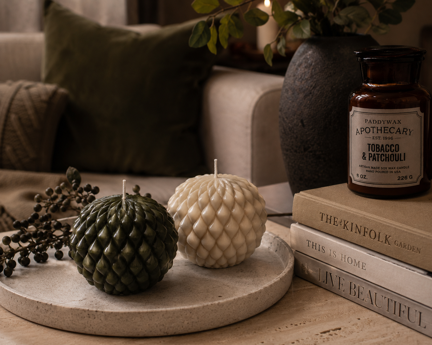 Moss and Cashmere Obsidian Bloom sculptural candles displayed on a stone tray beside a ceramic vase, books and apothecary jar in a warm interior setting.