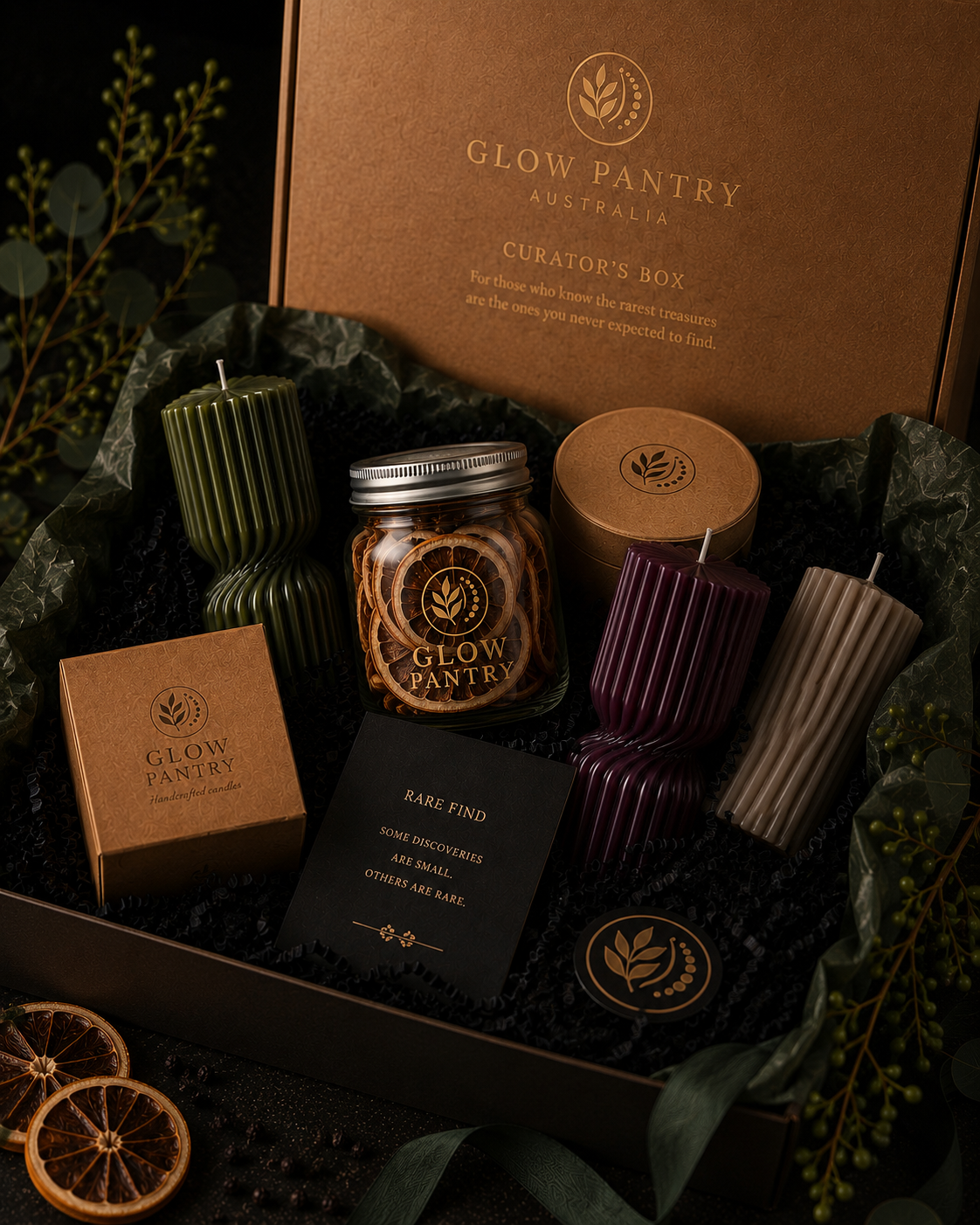 Glow Pantry Curator’s Box gift collection featuring a kraft presentation box, handcrafted sculptural candles in moss, mulberry and cashmere tones, a jar of dried citrus, luxury packaging and exclusive discovery items displayed on a dark botanical backdrop.