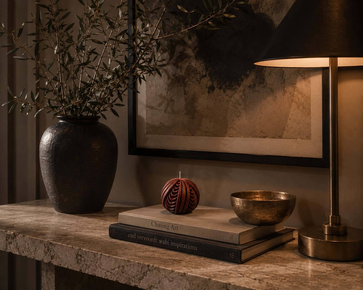 Deep terracotta Ember Obsidian Sphere candle styled on stacked books beside a ceramic vase and brass bowl, illuminated by warm lamp light in a moody contemporary interior.