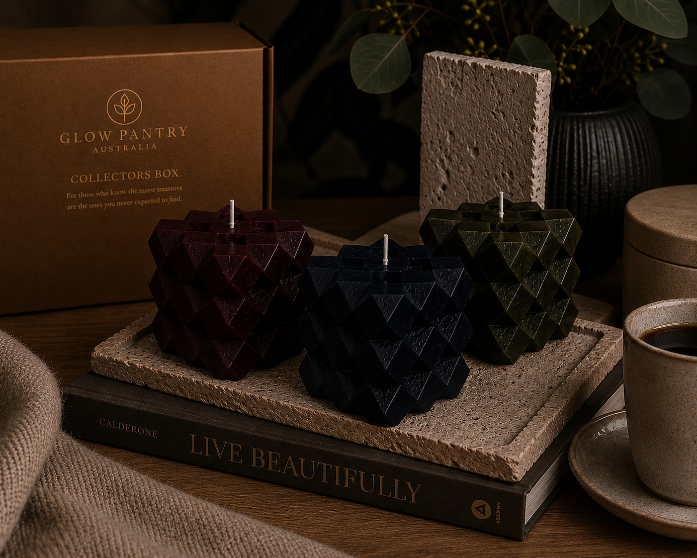 Discovery Cube sculptural candles in Ember, Midnight and Charcoal arranged on layered books and stone textures, with The Collector’s Box, ceramic vessels and botanical details creating a warm, curated interior scene.