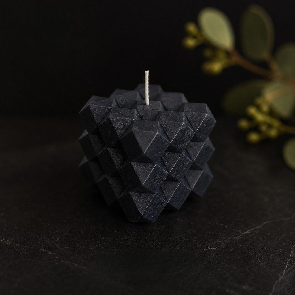 Midnight Discovery Cube sculptural candle in a rich charcoal-blue tone, photographed on a dark stone surface with eucalyptus foliage and berries softly blurred in the background. The faceted geometric form creates dramatic shadow and depth within a refined, low-light editorial setting.