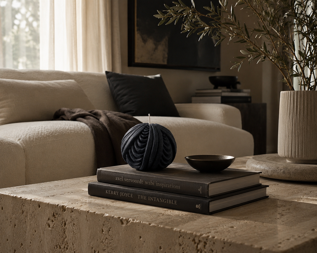 Midnight Obsidian Sphere sculptural candle styled atop stacked design books on a travertine coffee table, paired with a decorative bowl and olive branches in a warm contemporary living room.