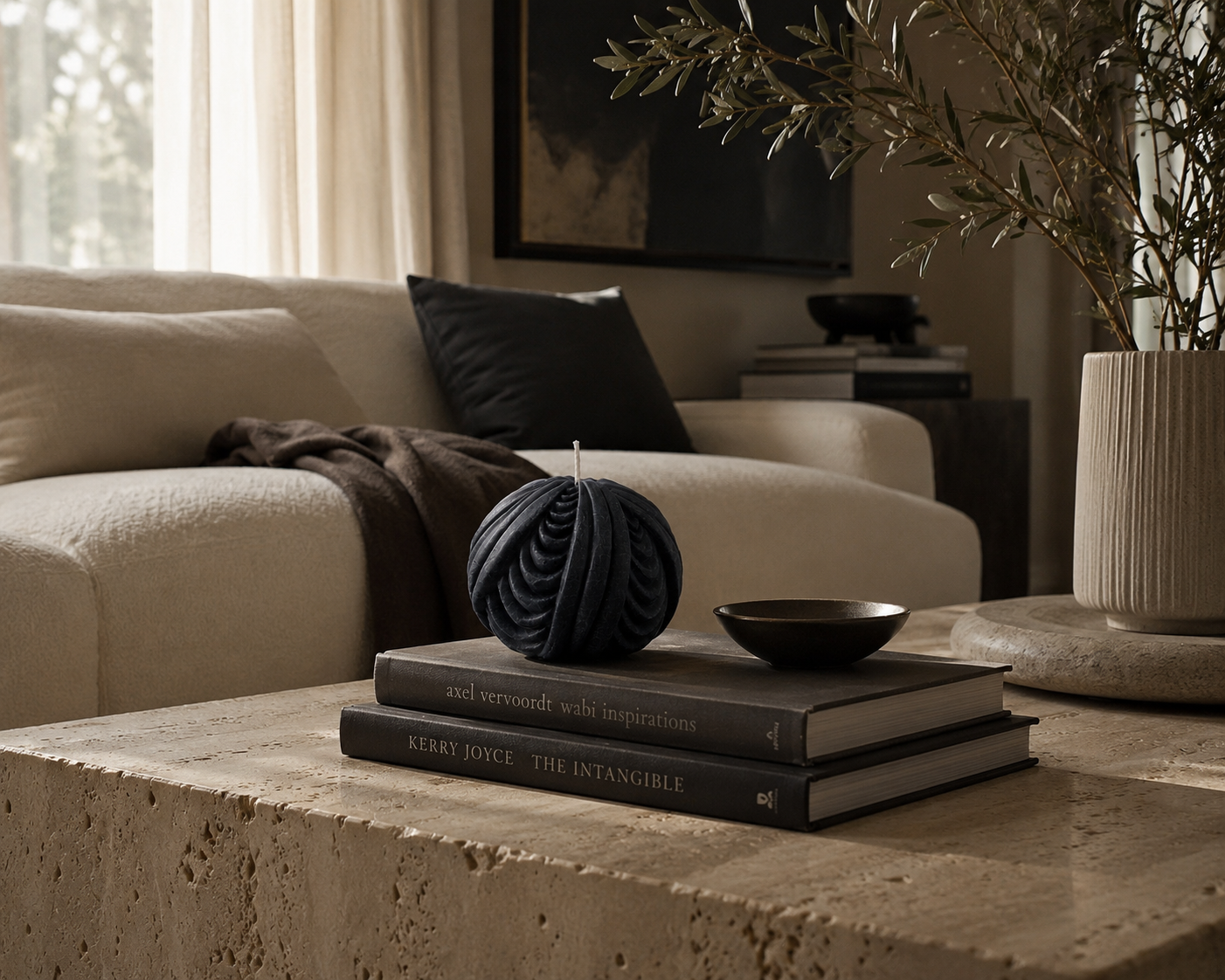 Midnight Obsidian Sphere sculptural candle styled atop stacked design books on a travertine coffee table, paired with a decorative bowl and olive branches in a warm contemporary living room.