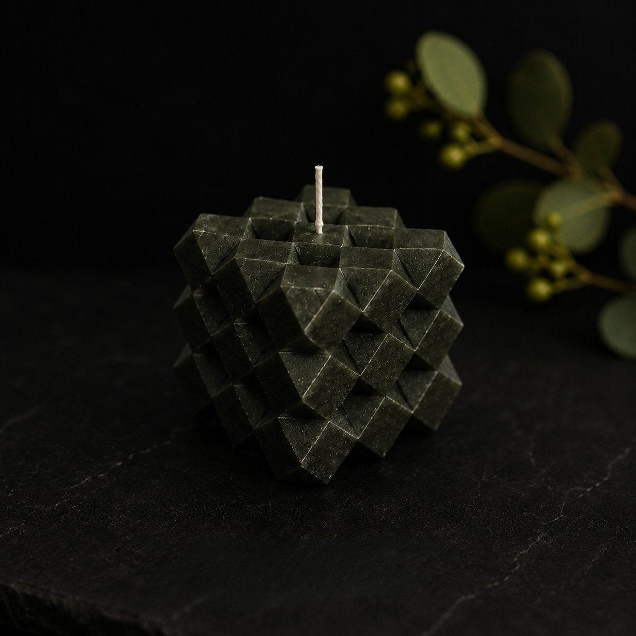 Moss Discovery Cube sculptural candle in a rich forest green tone, photographed on a dark stone surface with eucalyptus foliage and berries softly blurred in the background. The faceted geometric form creates depth and texture within a refined, low-light editorial setting.