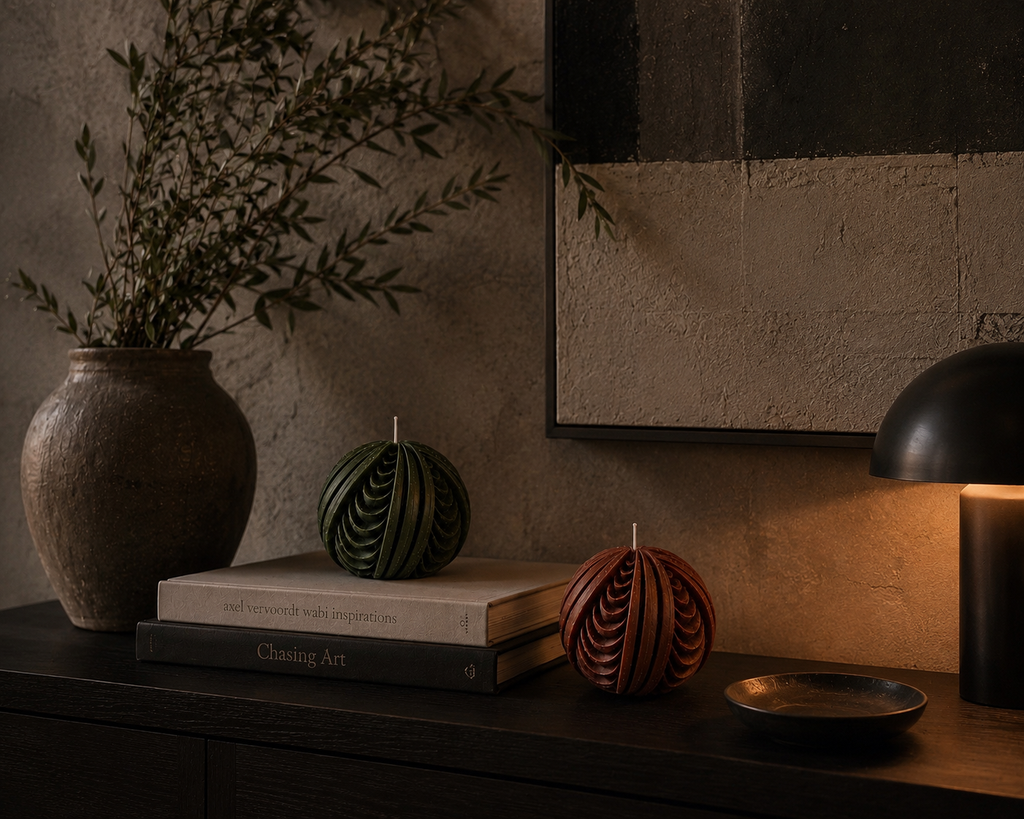 Glow Pantry Obsidian Sphere sculptural candles in Moss and Ember displayed on stacked books beside a ceramic vase and ambient lamp, styled in a warm, contemporary interior.