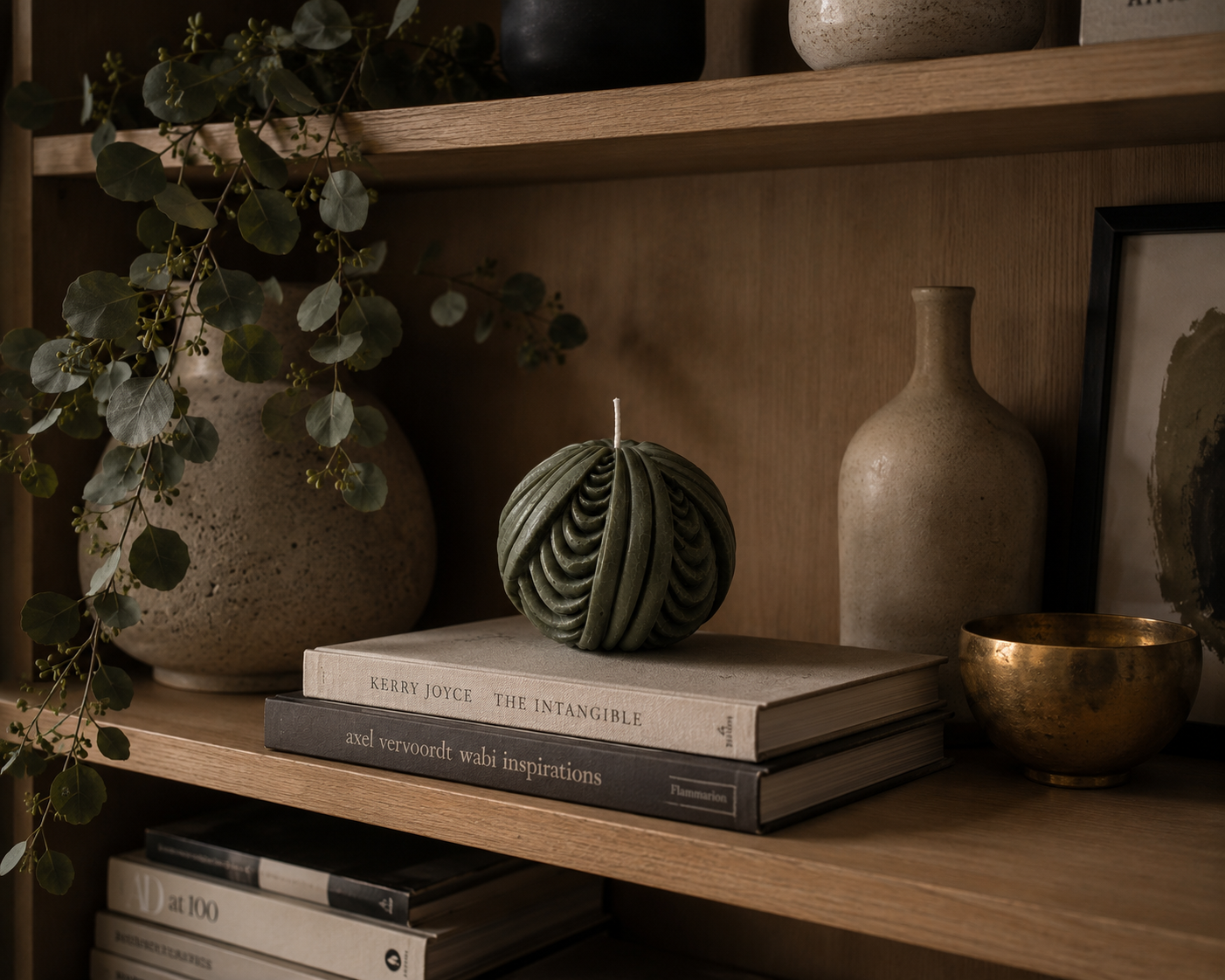 Deep moss-green Obsidian Sphere candle styled on stacked art books within a warm timber shelf display, surrounded by ceramic vessels, brass details and cascading eucalyptus branches.