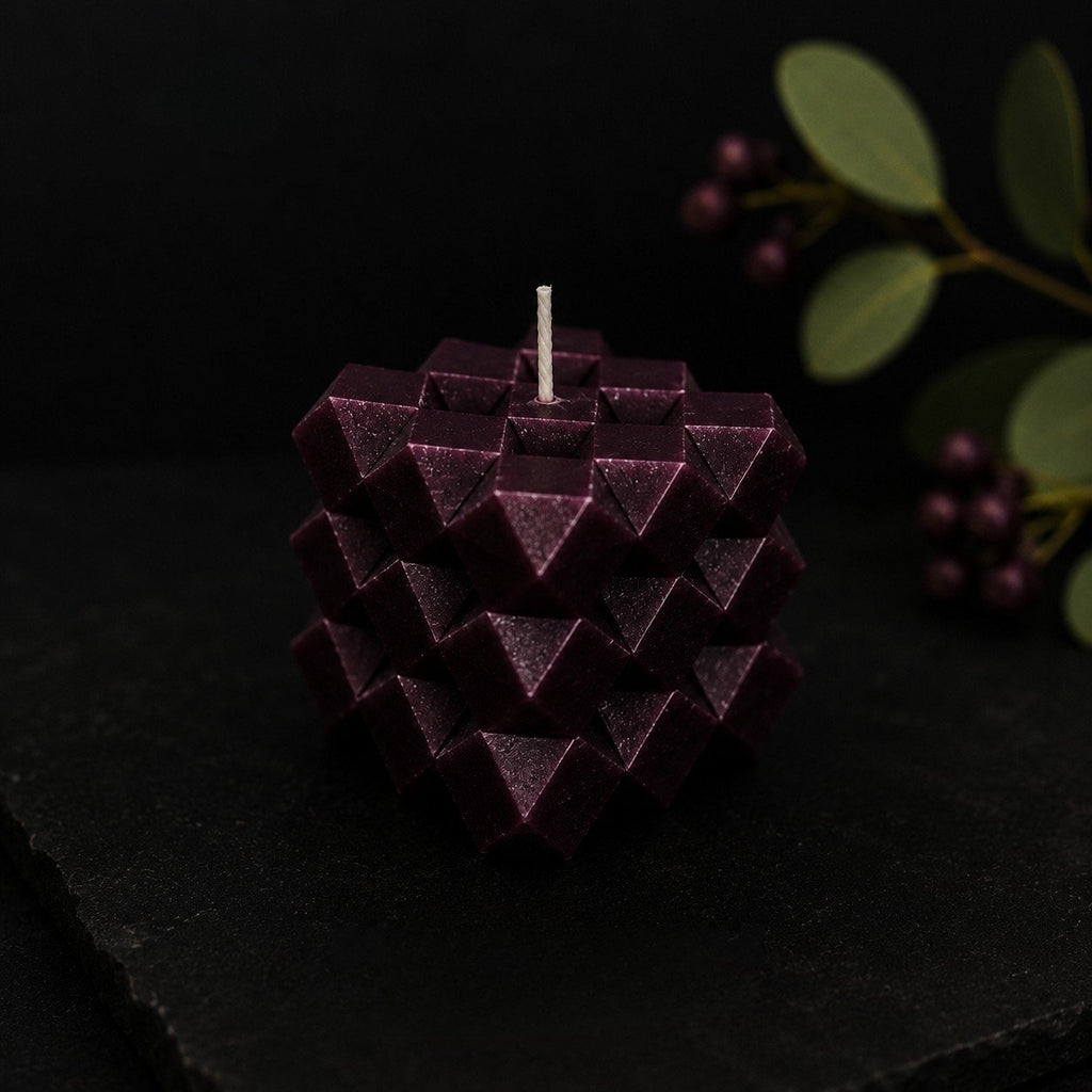 Mulberry Discovery Cube sculptural candle in a rich blackcurrant-plum tone, styled on a dark stone surface with soft eucalyptus foliage and berries in the background. Geometric faceted design with a moody editorial aesthetic.