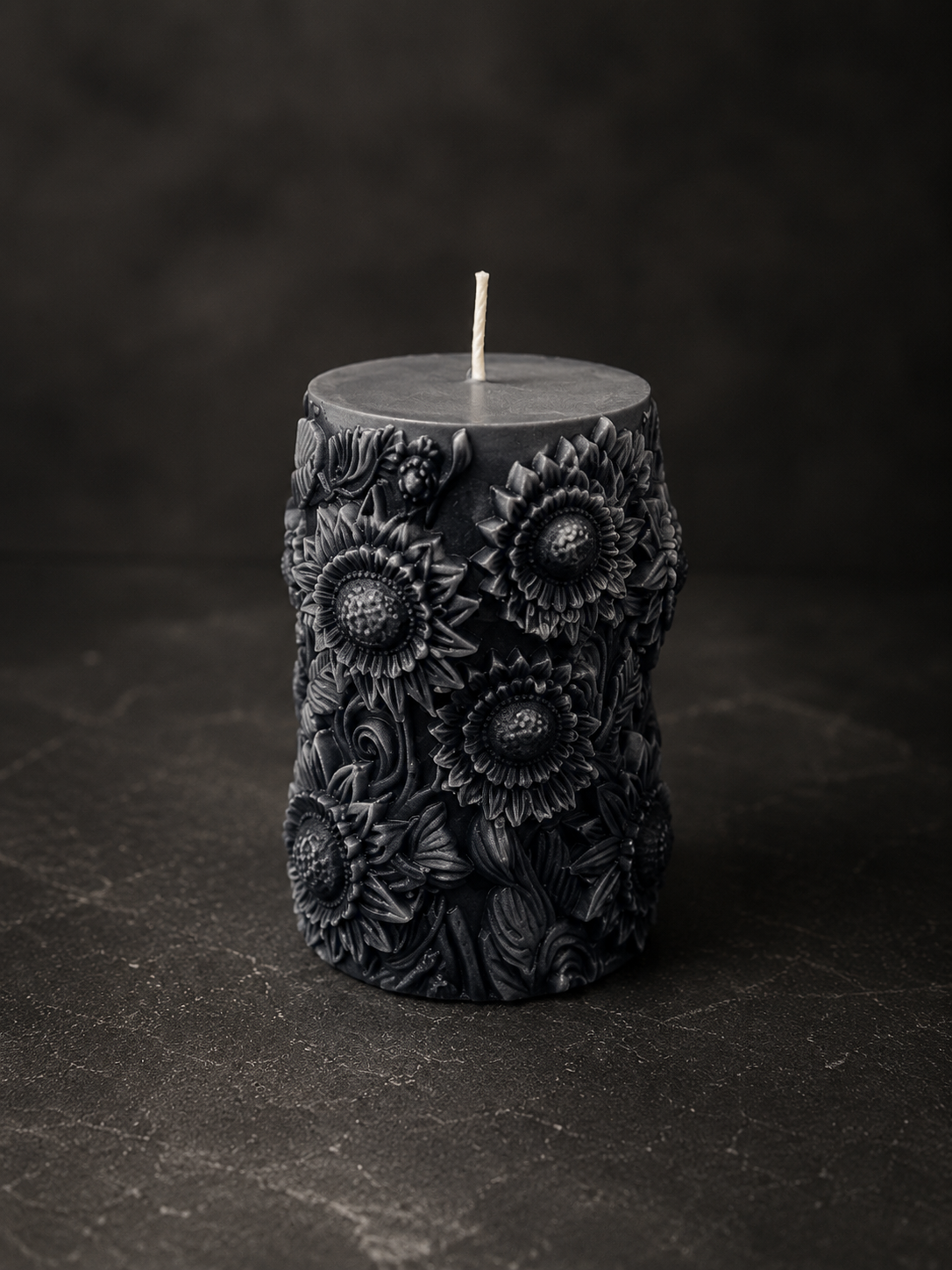 The Mourning Alchemist sculptural candle in layered charcoal and black tones with embossed sunflower detailing on a dark studio background.