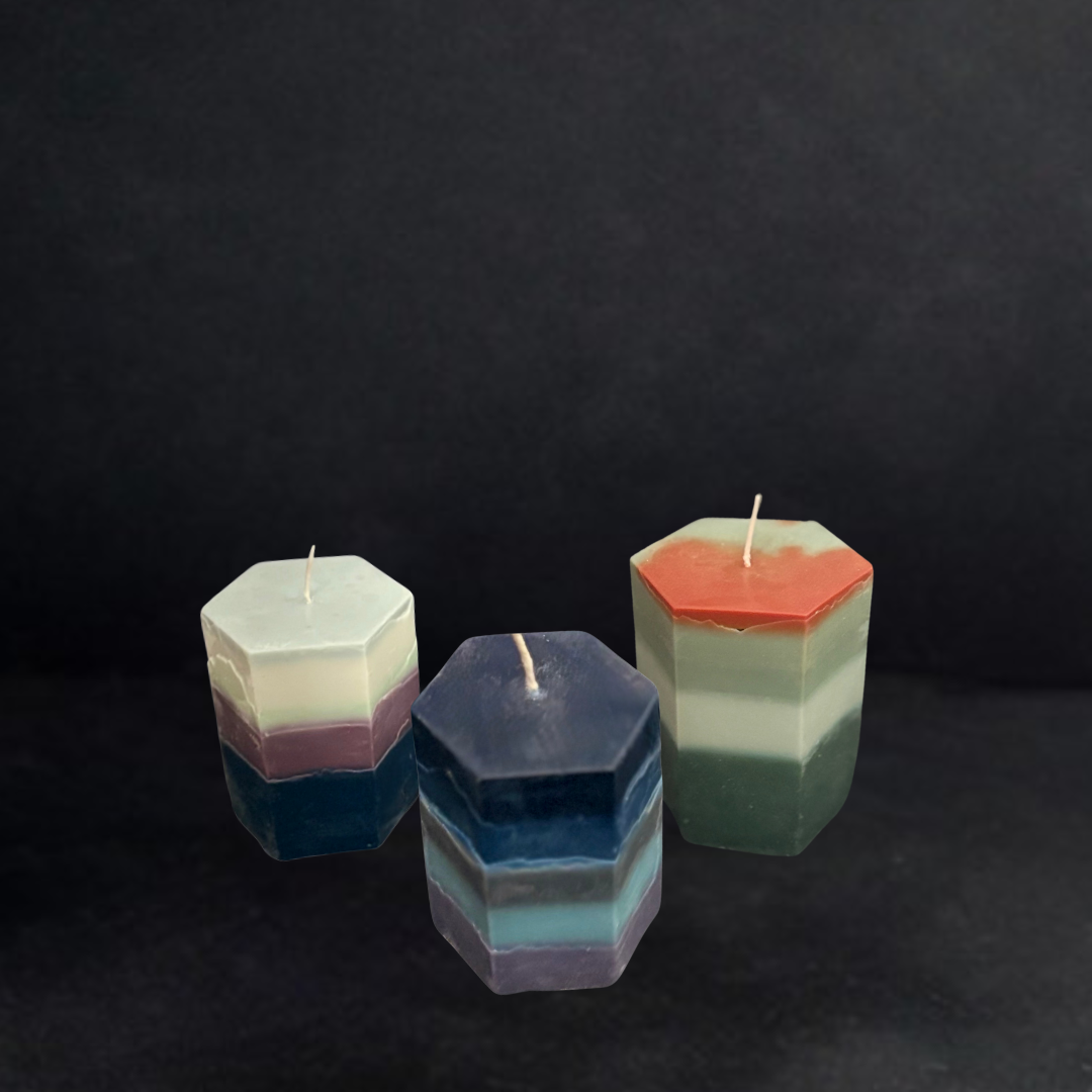 Three handcrafted hexagonal pillar candles with layered translucent pigments in blue, lilac, cream, and sage tones, styled against a dark studio backdrop.
